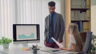 İş arkadaşıyla şirket büyüme göstergelerini tartışan erkek ekonomist ofis odasındaki bilgisayar ekranında grafiğin yanında duruyor.