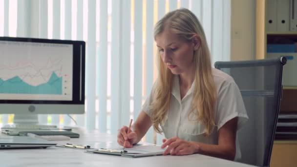 一个快乐的年轻女商人坐在写字台旁边 拿着商业图表 看着相机 一边做文书工作的画像 — 图库视频影像