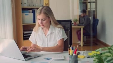 Yoğunlaşmış kadın laptopta iş raporu hazırlarken iş arkadaşı arka planda geniş modern ofiste oturuyor.