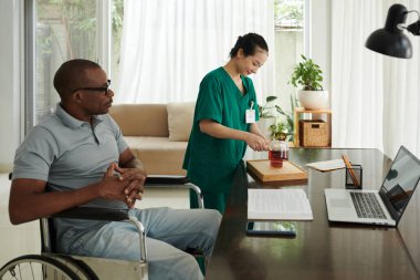 Tekerlekli sandalyede oturan engelli için çay yapan sosyal hizmet görevlisi.