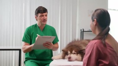 Yeşil üniformalı bir erkek veterinerin tıbbi karttaki hasta köpek hakkında notlar yazarken sahibinin pofuduk evcil hayvanının yanında oturduğu orta boy bir fotoğraf.