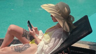 Tatilci kadın internet şakalarına akıllı telefondan gülerken dinlenme odasında havuzun yanında uzanır.