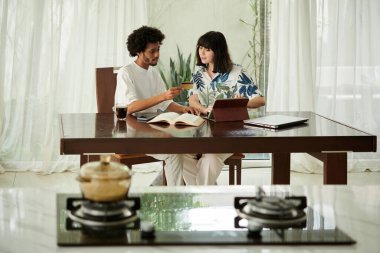 Genç esmer kadın banka bilgilerini girerken kocasının elinde tuttuğu kredi kartına bakıyor.