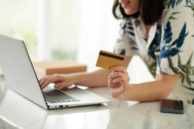 Kredi kartı tutan ve bilgisayarın klavyesinin tuşlarına basan genç bir kadın tarafından çevrimiçi dükkanda seçilen ürünler için ödeme yapılıyor.