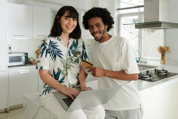 Young smiling husband and wife in casualwear looking at camera in the kitchen while making online order and paying by credit card for goods