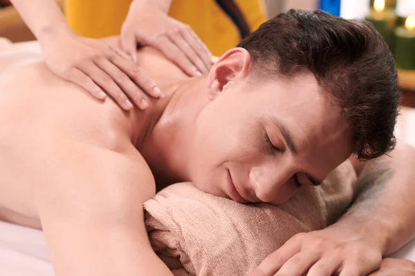 Young pleased man lying on his belly on couch and getting professional spa massage of back and shoulders by masseuse working in salon