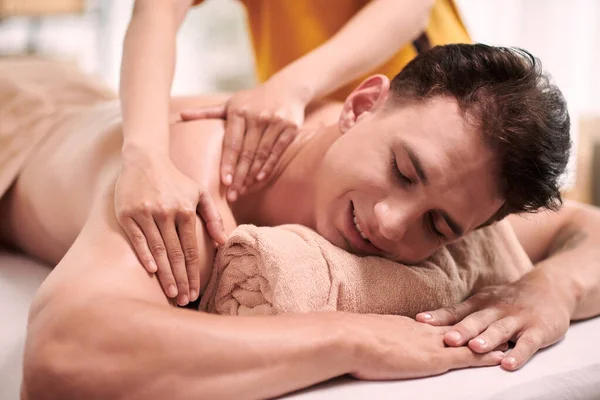 Happy young man keeping chin on rolled soft towel while relaxing on couch during procedure of professional spa massage in beauty salon