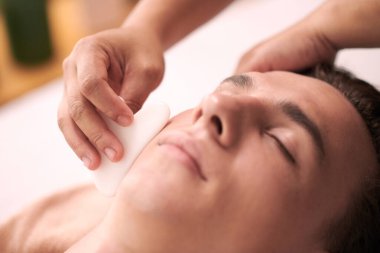 Kozmetik uzmanı, insanın yüzüne masaj yaparken gua sha kullanıyor.
