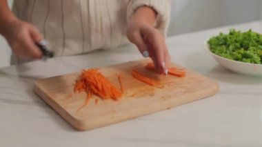 Kadın ellerinin ahşap tahtada havuç dilimlemesi ve mutfakta akşam yemeği pişirirken domatesli ve yeşillikli kase eklemesi.