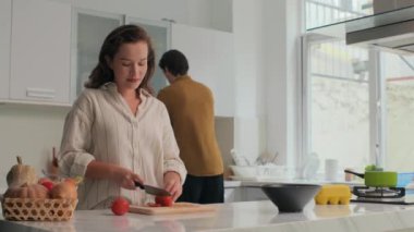 Genç çift mutfakta birlikte yemek pişiriyor, kadın salata için sebze keserken erkek de modern mutfakta ona yemek veriyor.