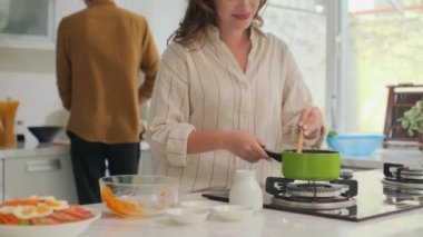 Eş mutfakta yemek pişirirken, genç kadın tuz çorbası içerken erkekler salata keserken.