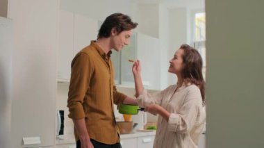 Modern mutfakta birlikte yemek pişirirken erkek arkadaşına bir kaşık çorba veren orta boy bir kız resmi.