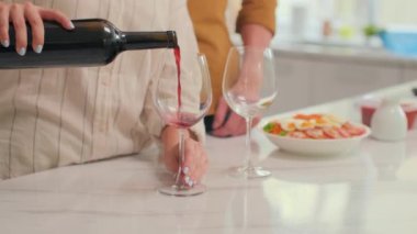 Romantik bir akşam yemeğinden önce erkek arkadaşı yanında dururken kırmızı şarap dolduran bir kadın resmi.