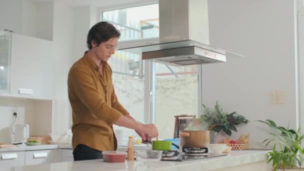 中长镜头的白人年轻人 一边用勺子尝橙汤 一边在厨房的炉灶上用绿色平底锅煮 — 图库视频影像
