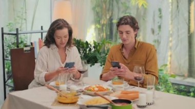 Evde romantik bir akşam yemeği yerken sosyal medyada arkadaşlarına mesaj atan iki genç adam ve kadının orta boy fotoğrafı.