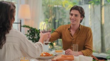 Evde romantik bir akşam yemeği sırasında genç eşlerin birbiriyle konuştuğu ve kırmızı şarapla kadeh tokuşturduğu orta boy bir fotoğraf.