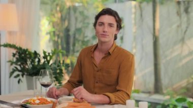 Beyaz bir adamın portresi. Yemek masasında oturmuş, kırmızı şarap içiyor ve kameraya bakıyor.