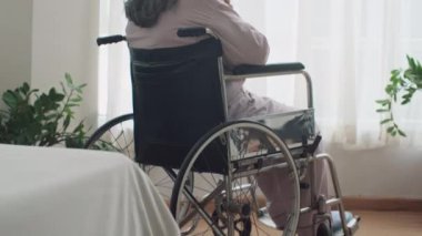 Tekerlekli sandalyedeki kadın emeklinin huzurevinde pencerenin yanında tek başına oturması.