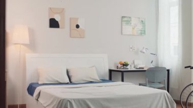 Büyük boy yatağı, yazı masası, duvarda resimleri ve pastel renkli tekerlekli sandalyesi olan engelli insanların orta boy uzun yatak odası.