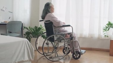 Tekerlekli sandalyedeki üzgün olgun kadının yatak odasında vakit geçirmesi ve pencereye bakması.