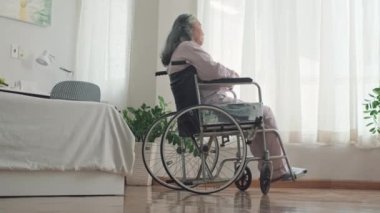 Özürlü yaşlı bir kadının bakımevinin yatak odasında, pencerenin yanında oturmasının düşük açılı görüntüsü.