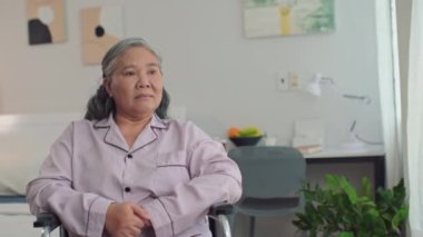 Yatak odasındaki tekerlekli sandalyede kameraya bakan yaşlı hasta kadının portresi.
