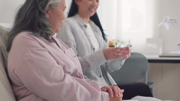 若いアジアの娘は病気の高齢の母親の世話をし 看護の家を訪問するときに水のグラスを与えます — ストック動画