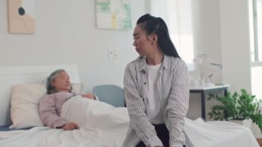 Orta boylu, ağlayan Asyalı bir kız çocuğunun ciddi şekilde hasta olan yaşlı annesinin yatağında huzurevinde oturması.