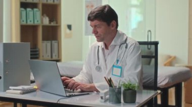 Klinikteki masada oturup kameraya bakarken dizüstü bilgisayardaki gülümseyen doktorun portresi.