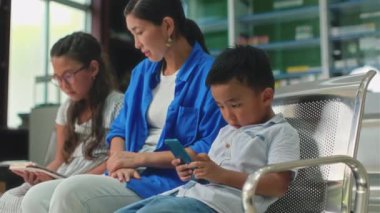Anne ve iki çocuğun klinikte muayeneyi beklerken dijital cihazlar kullanırken orta uzunlukta bir görüntüsü.