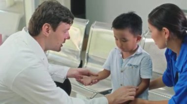 Kafkas doktorunun klinikte otururken küçük bir hasta ve annesiyle kol ağrısı hakkında konuşurken orta boy fotoğrafı.