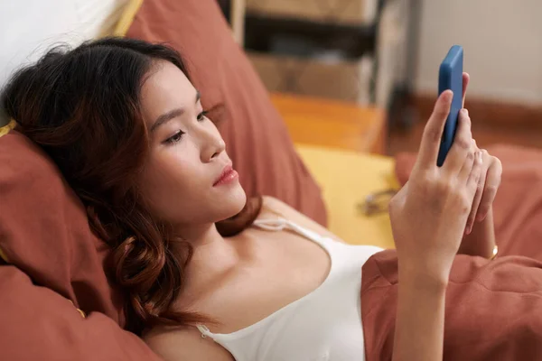 Young woman resting in bed, checking social media on smartphone before sleep