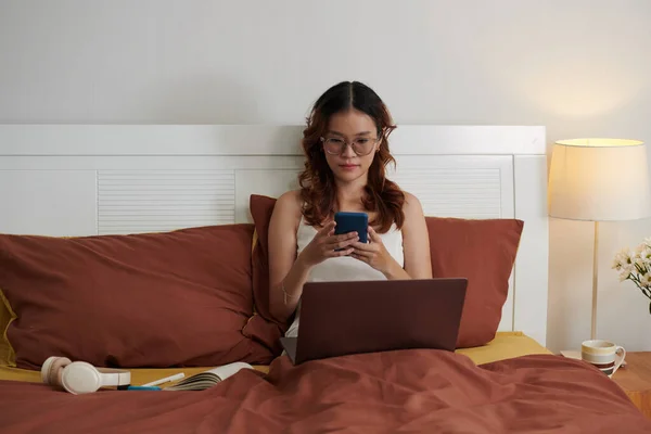 Young woman sitting in bed with laptop and texting coworkers