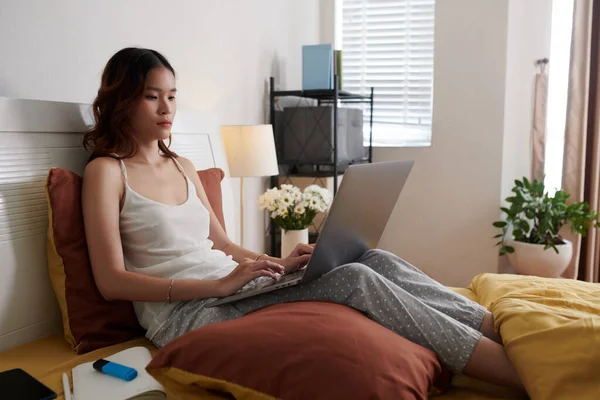 Serious young woman in pajamas sitting on bed and working on laptop