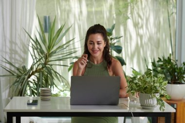 Evde dizüstü bilgisayarda çalışan gülümseyen genç bir kadın. Etrafta bir sürü bitki var.