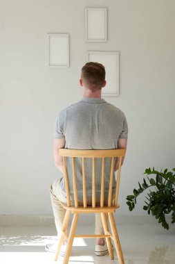Genç adam sandalyede oturuyor ve duvarda asılı boş çerçevelere bakıyor.