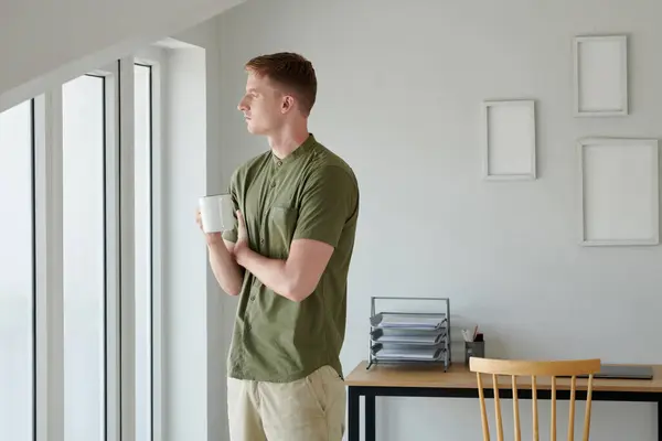 一个多愁善感的男人 拿着一杯咖啡从公寓窗户往外看 — 图库照片