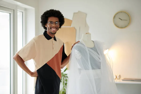 Smiling Black fashion designer standing next to mannequin with draped fabric