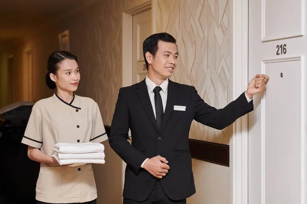 Smiling hotel manager knocking in door, maid with fresh towels standing next to him