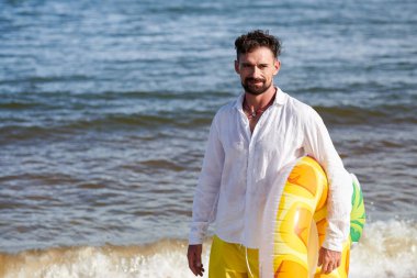 Yüzme yüzüğüyle sahilde duran gülümseyen adamın portresi.