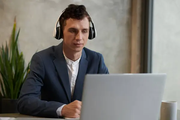 笑顔の若いビジネスマンは ノートパソコンの画面でレポートを読んでヘッドフォンで音楽を聞いています — ストック写真