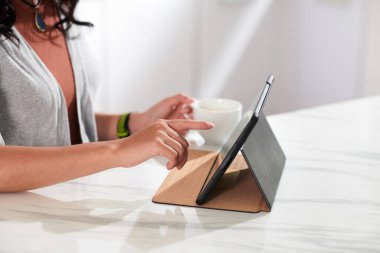 Dijital tablet kullanan ve masada kahve içen genç bir kadının çevrimiçi video izlediği yakın plan.
