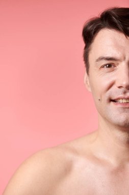 Pembe arka planda duran pozitif tişörtsüz olgun bir adamın portresi.