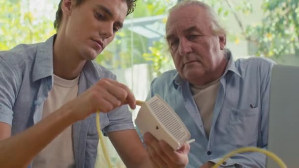 Сбитый Молодой Человек Помогал Дедушке Подключить Роутер Ноутбуку Сидя Партой — стоковое видео