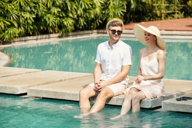 Genç çift yüzme havuzunun yanında oturuyor ve konuşuyor, tatil köyünde vakit geçiriyorlar.