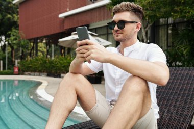 Güneş gözlüklü genç adam açık havuz kenarında güneşli yatakta oturuyor ve cep telefonunu internet iletişimi için kullanıyor.