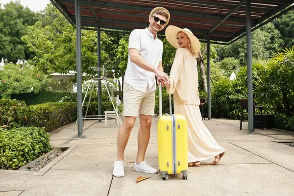 Portrait of young couple in hats with luggage smiling at camera standing outdoors