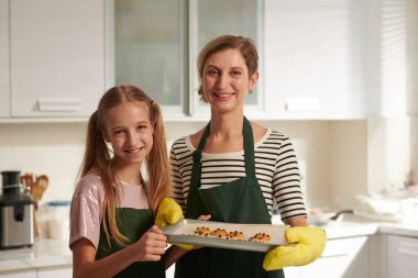 Gülümseyen anne ve genç kızının birlikte pişirdikleri kurabiyelerle tepsiyi tutarken portresi.