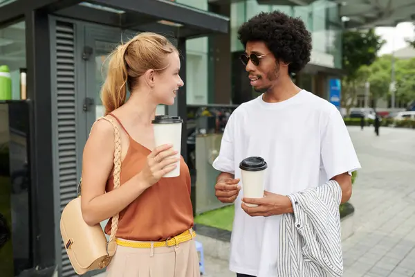 Positive young colleagues standing in the street, drinking coffee and discussing news