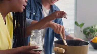 Çok ırklı bir çiftin evde yemek pişirirken tencereye un kattığı orta boy bir fotoğraf.